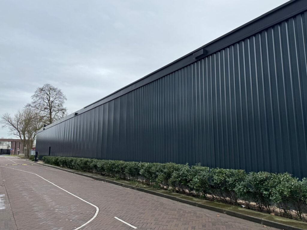 Lang industrieel gebouw met zwarte metalen gevel en groene struiken.