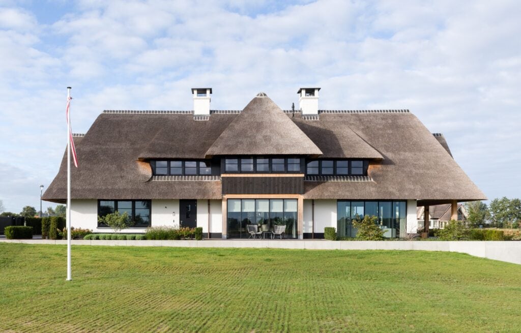 Luxe houten landhuis met rieten dak en grote ramen, omgeven door groene tuin.