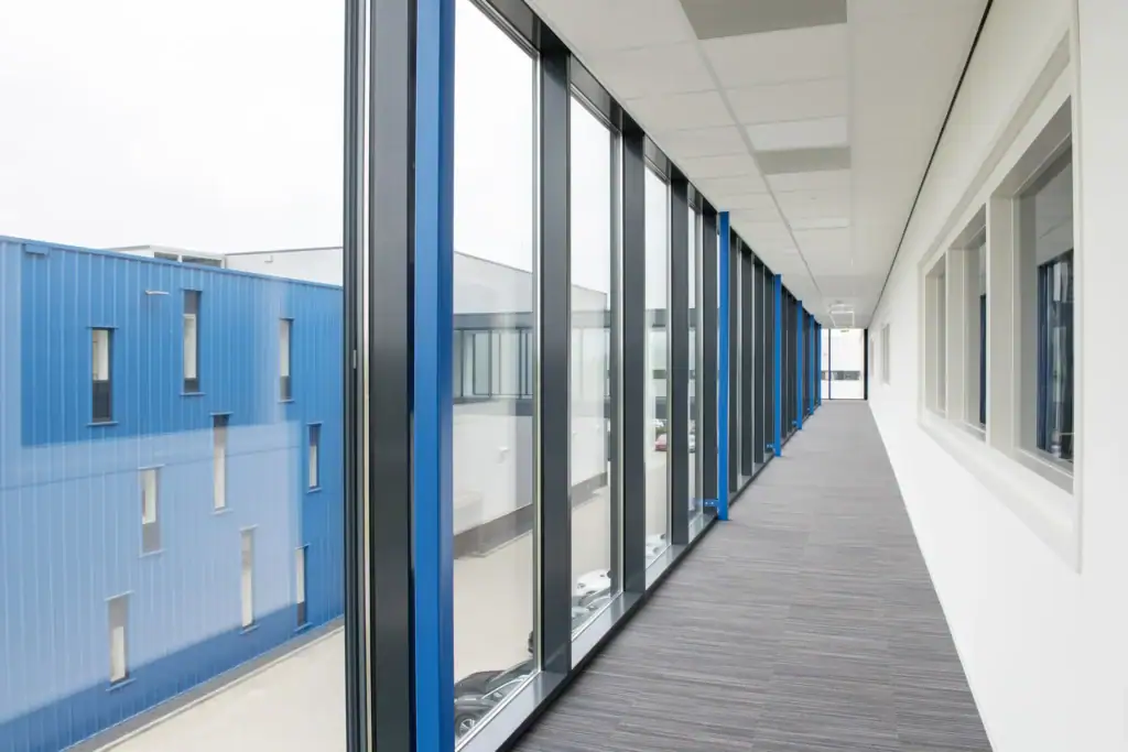 Lange glazen corridor in een modern kantoorpand met uitzicht op blauwe gebouwen.